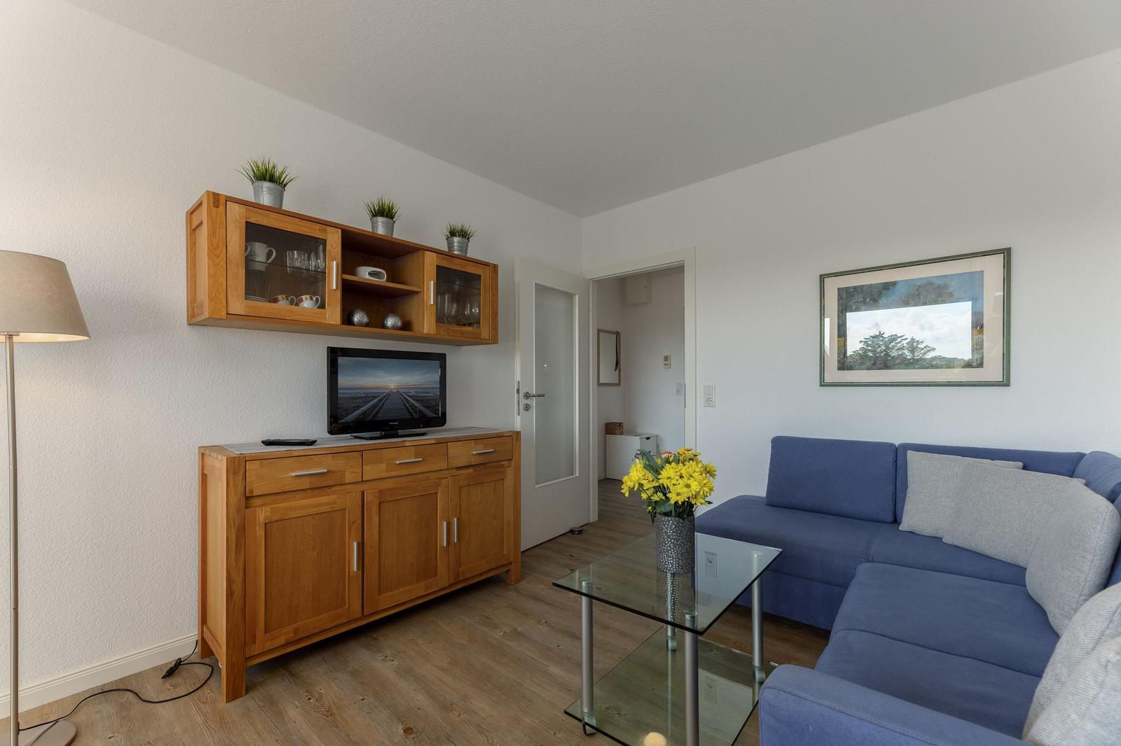 Wohnzimmer mit blauem Sofa, Glas-Tisch und Holzmöbeln. Fernseher und Pflanzen sind sichtbar.