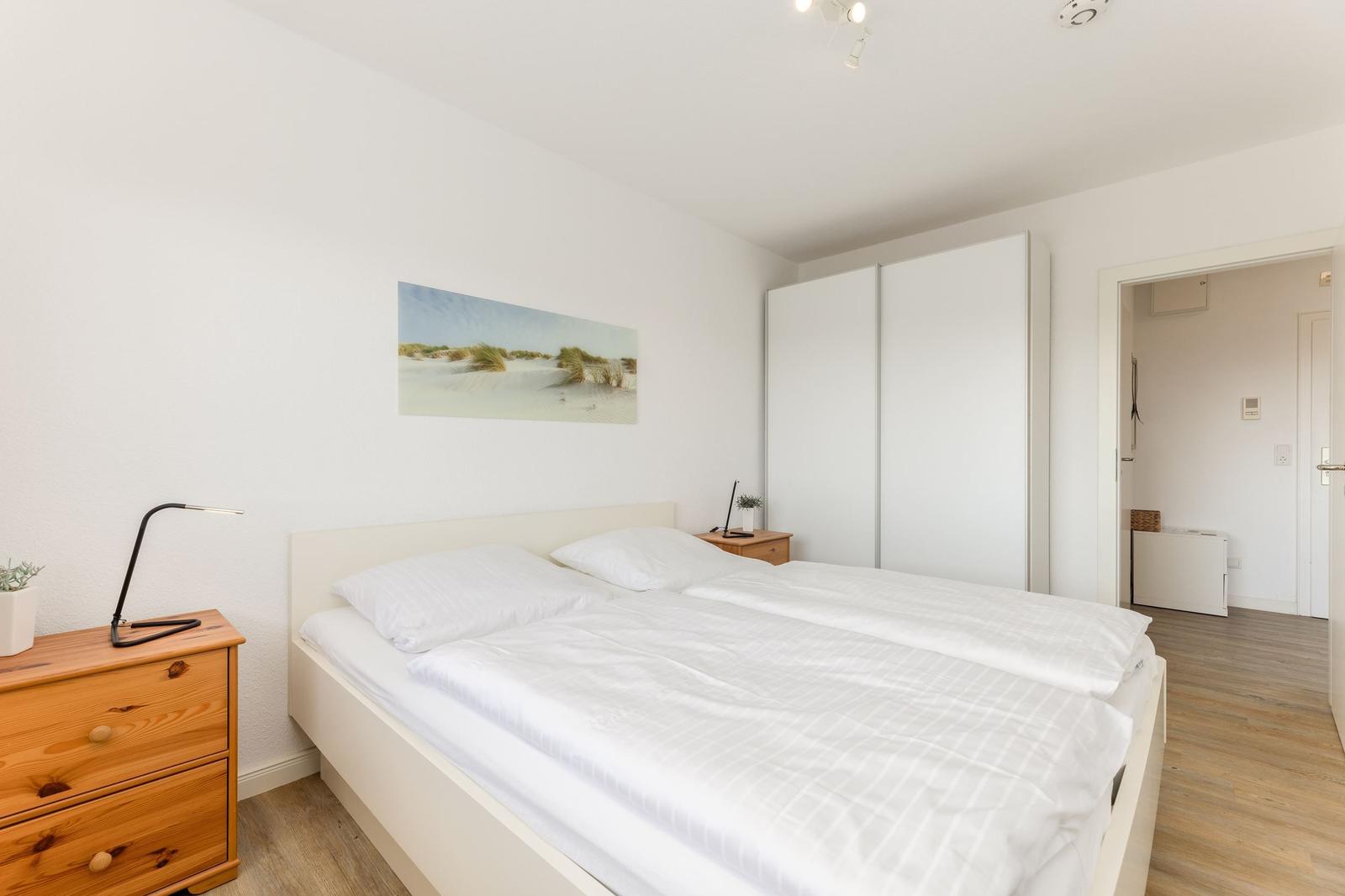 Schlafzimmer mit Doppelbett, Schrank und Nachttisch. Wandbild zeigt Dünenlandschaft.