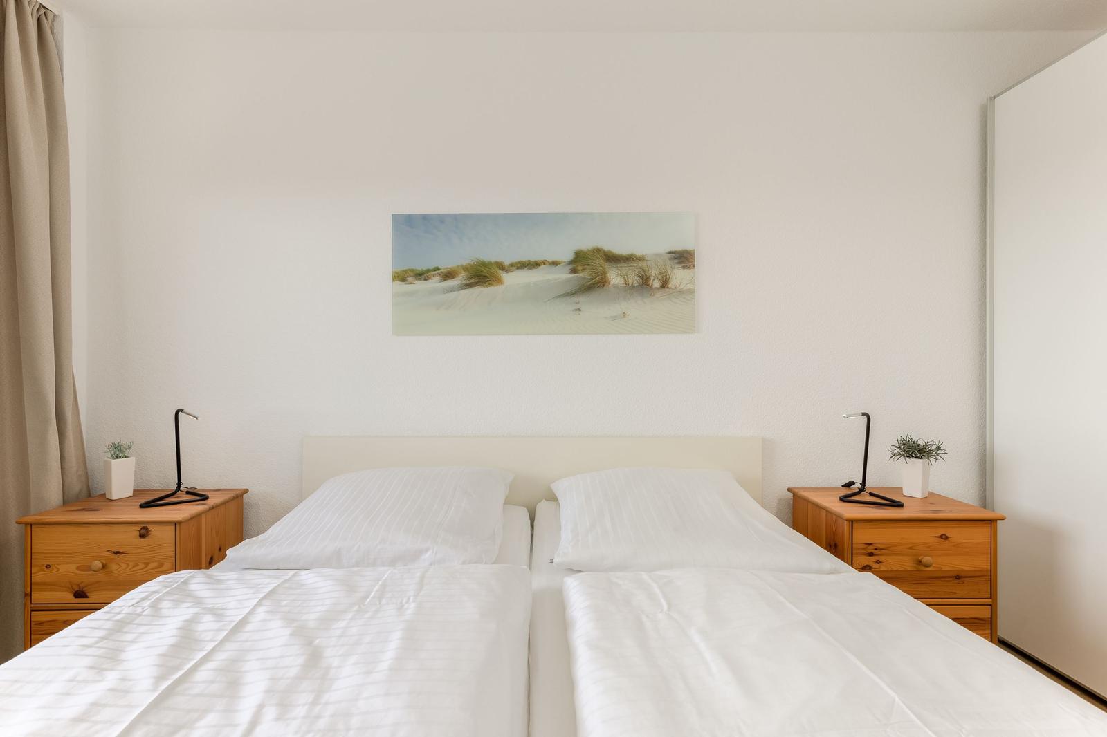 Zwei Betten mit weißen Bettwäsche, Holznachttische und Wandbild mit Dünenmotiv.