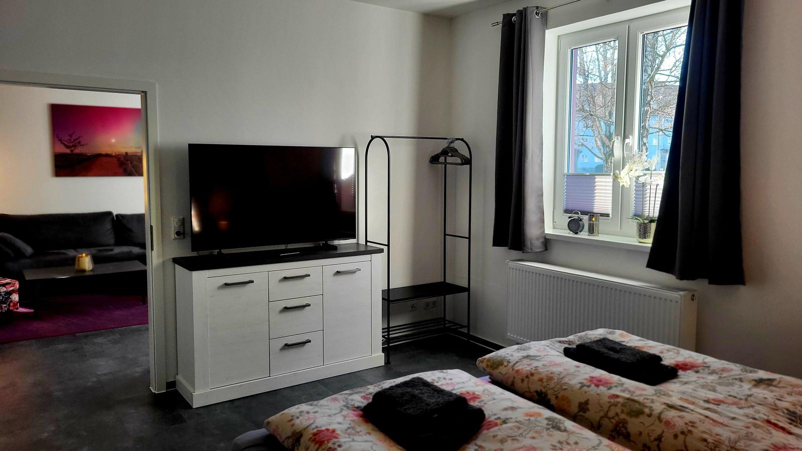 Zwei Betten, Fernseher und Kleiderständer im Schlafzimmer. Durchgang ins Wohnzimmer.