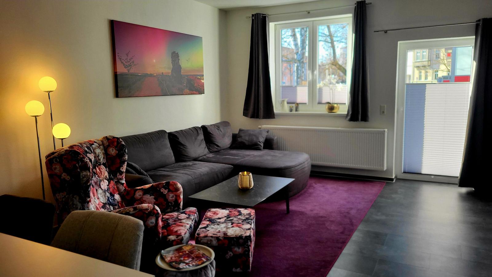 Wohnzimmer mit Sofa, Sessel und Teppich. Fenster und Tür mit Vorhängen.