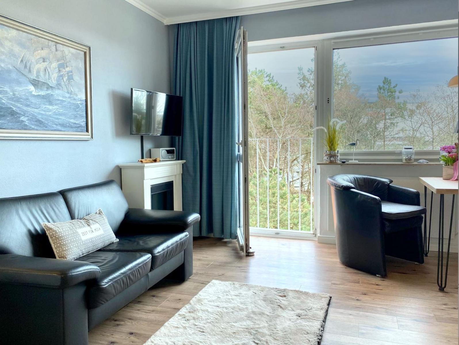 Wohnzimmer mit Sofa, Sessel und Balkonblick. Fernseher und Wandbild sind vorhanden.