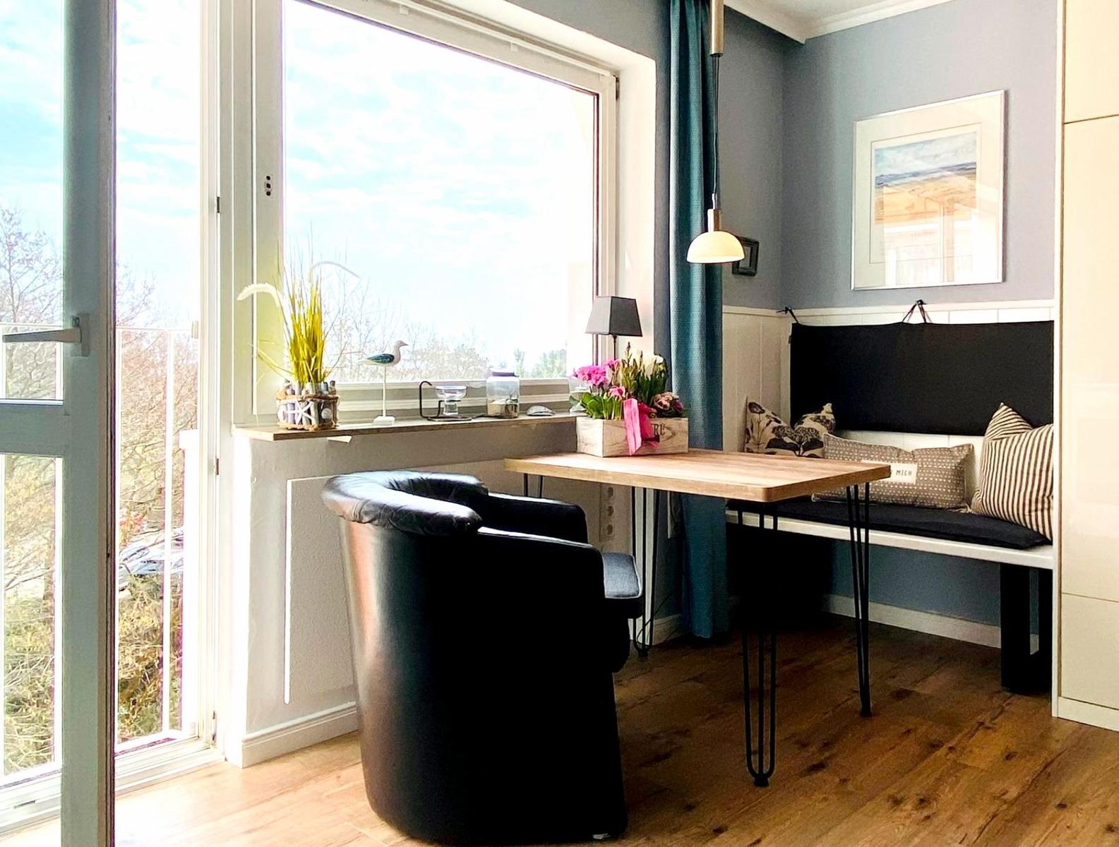 Ein Raum mit Tisch, Sitzbank und Stuhl. Durch Fenster und Tür ist Außenbereich sichtbar.