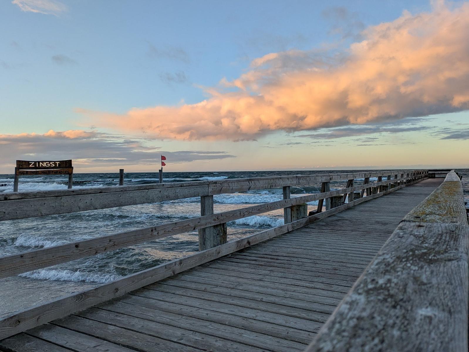 Holzbrücke führt zum Strand von Zingst. Im Hintergrund sind Wellen und Wolken zu sehen.