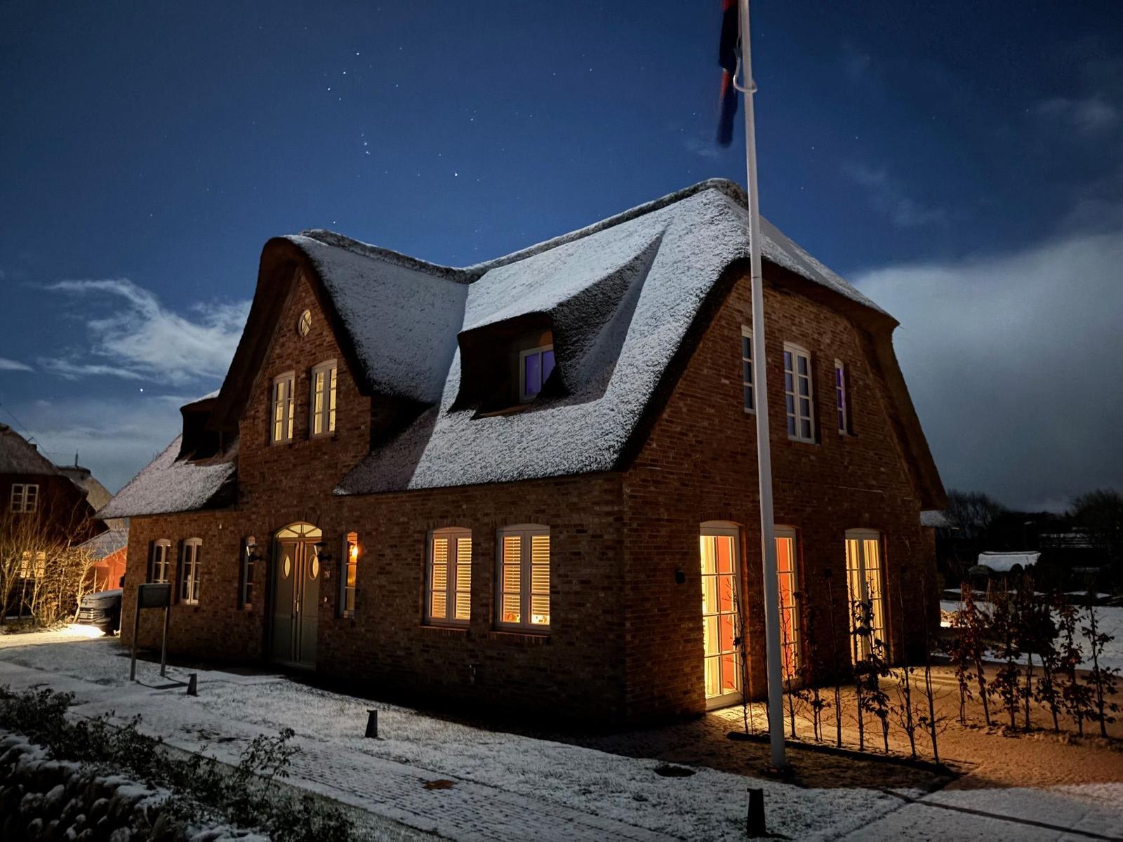 Schneebedecktes Haus mit erleuchteten Fenstern unter sternenklarem Himmel.