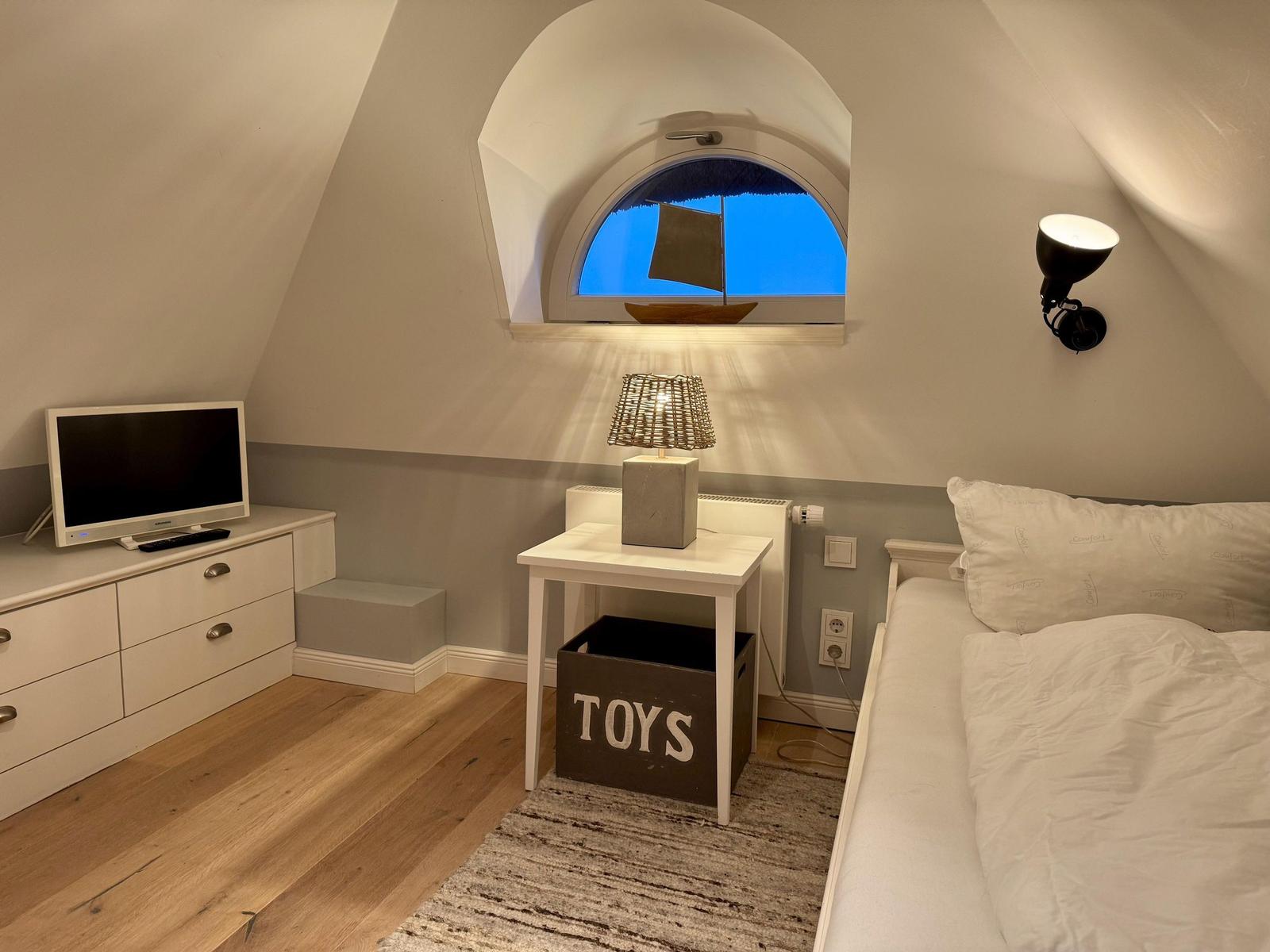 Schlafzimmer mit Dachschrägen, Bett, Nachttisch, Fernseher und Spielzeugkiste.