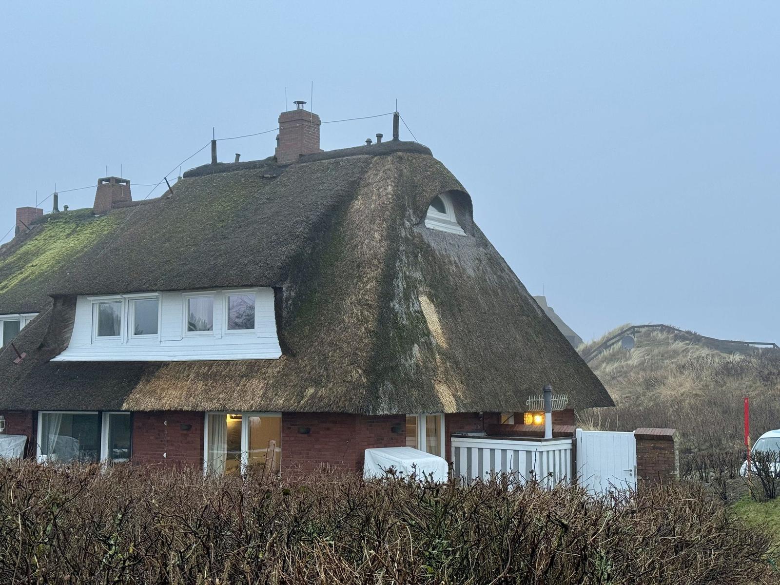 Dorfhaus mit strohgedecktem Dach und Fenstern. Hinterhof mit Hecke und Gartenmöbeln.