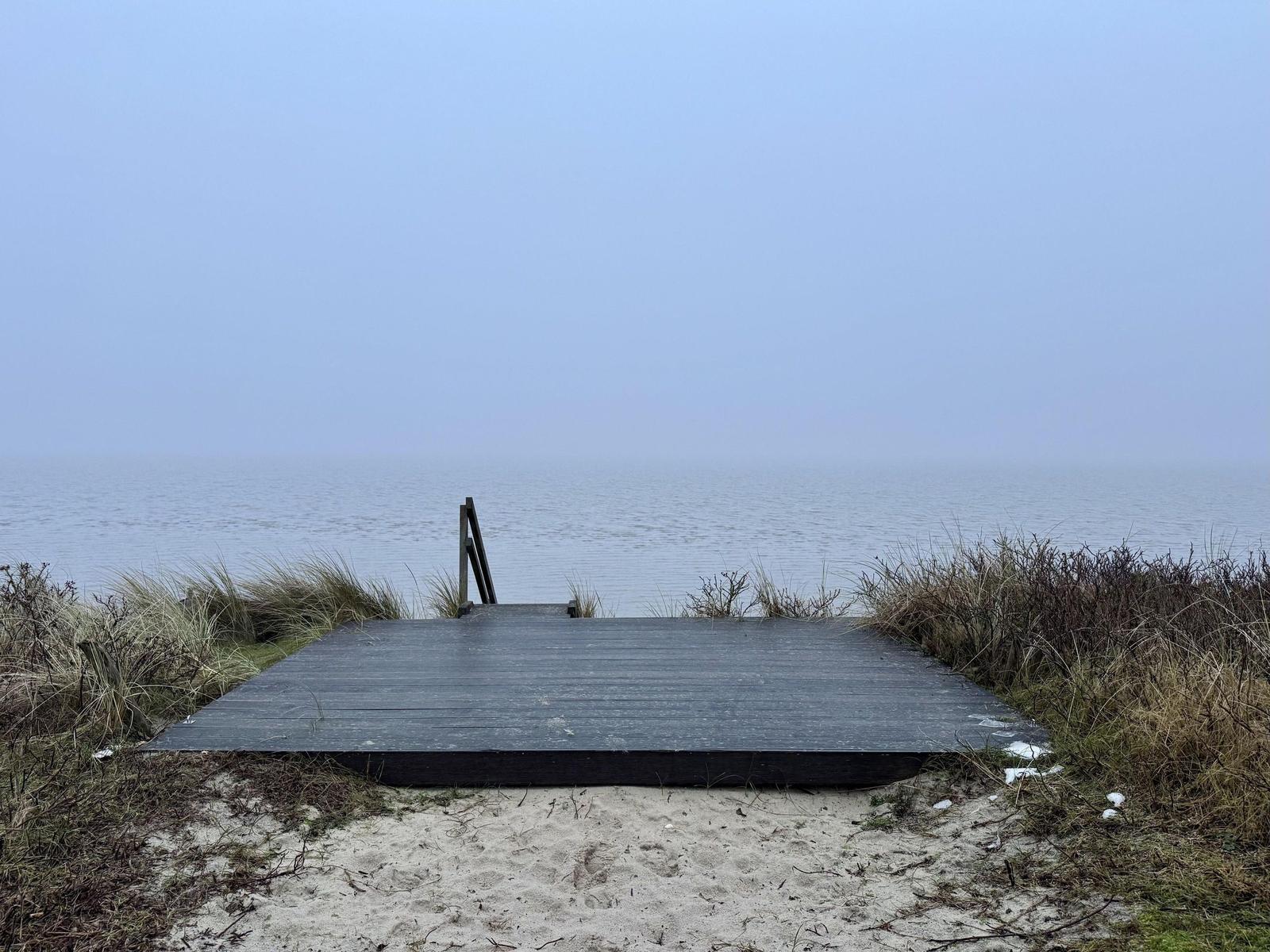 Holzterrasse führt zum Wasser. Nebel umgibt See und Strand.