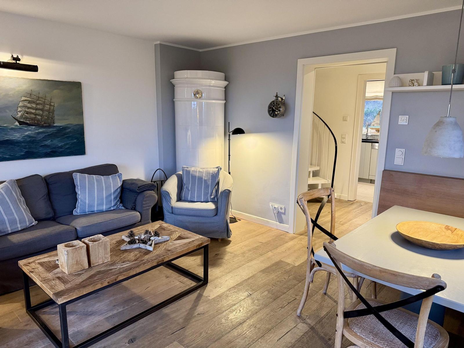 Wohn- und Essbereich mit Sofa, Sessel und Holztisch. Weiße Wanduhr und Kamin sichtbar.