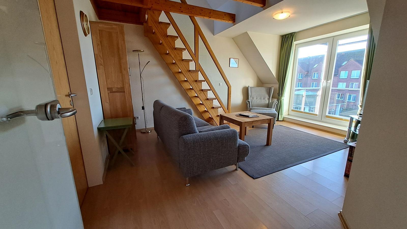 Wohnzimmer mit Sofa, Tisch und Holztreppe. Große Fenster und Holzboden.