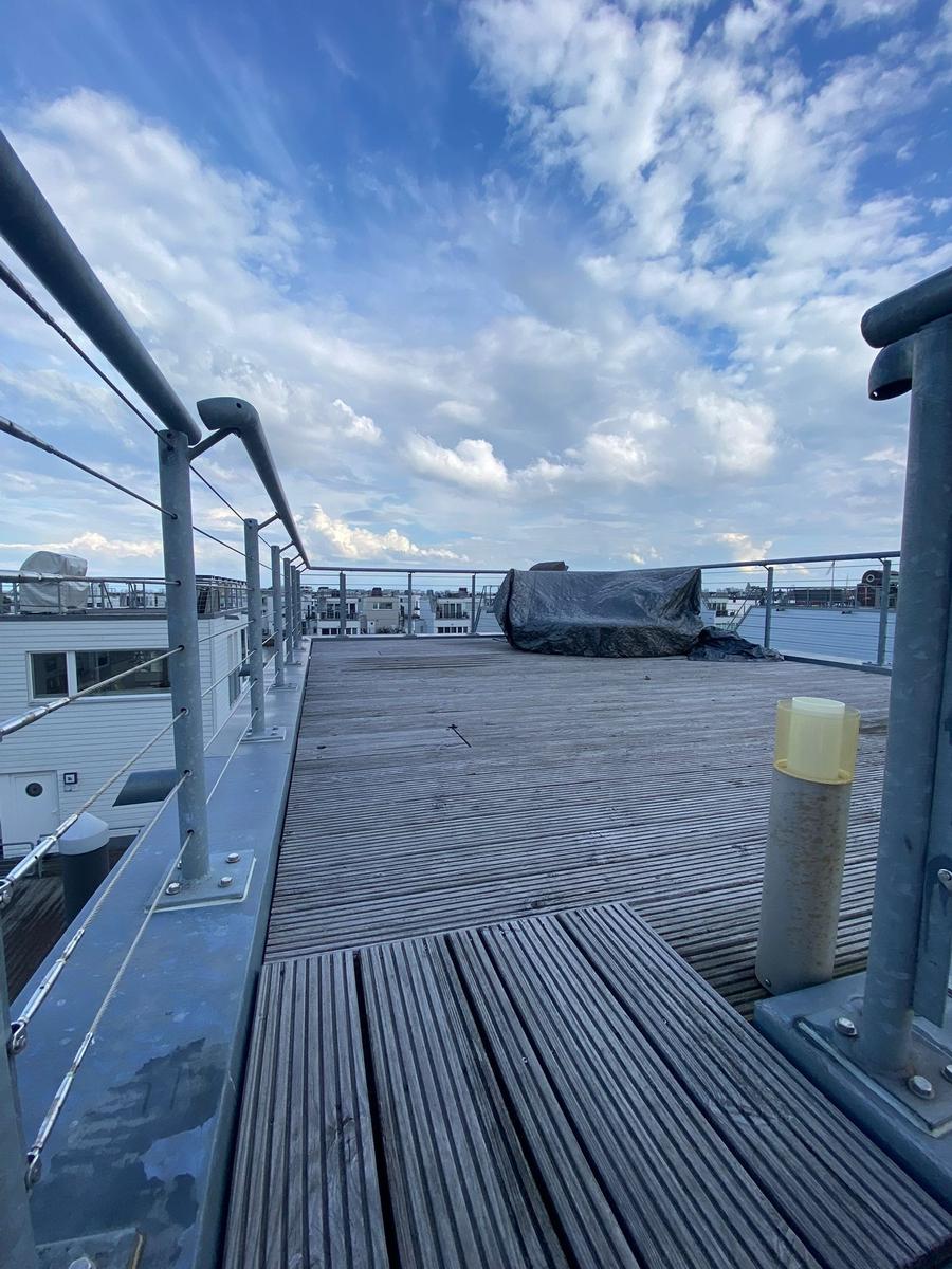 Roof terrace with wooden floor and metal railing. Buildings and sky with clouds are visible in the background.
