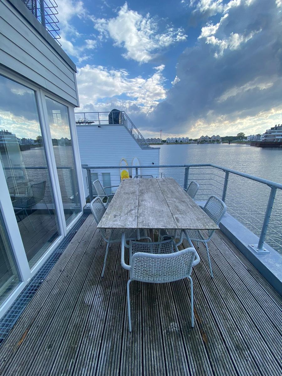 Deck with table and chairs by the water. View of harbor and buildings across the water.