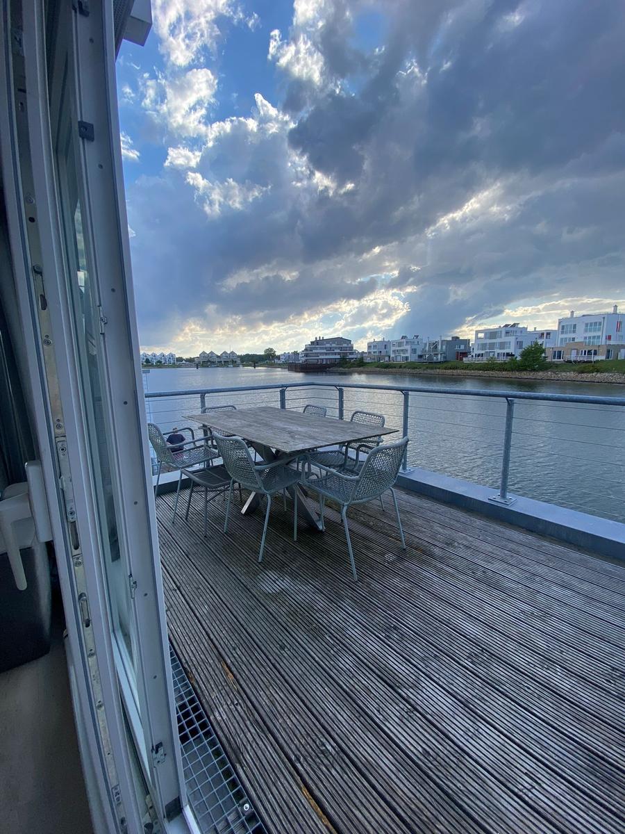 Terrace with table and chairs by water, overlooking houses and cloudy sky.