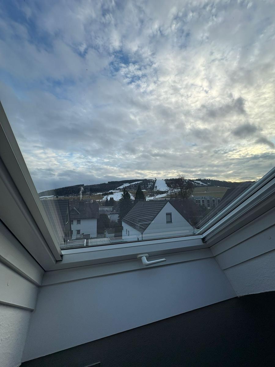 Durch das Fenster blickt man auf eine verschneite Landschaft mit Häusern und bewölktem Himmel.