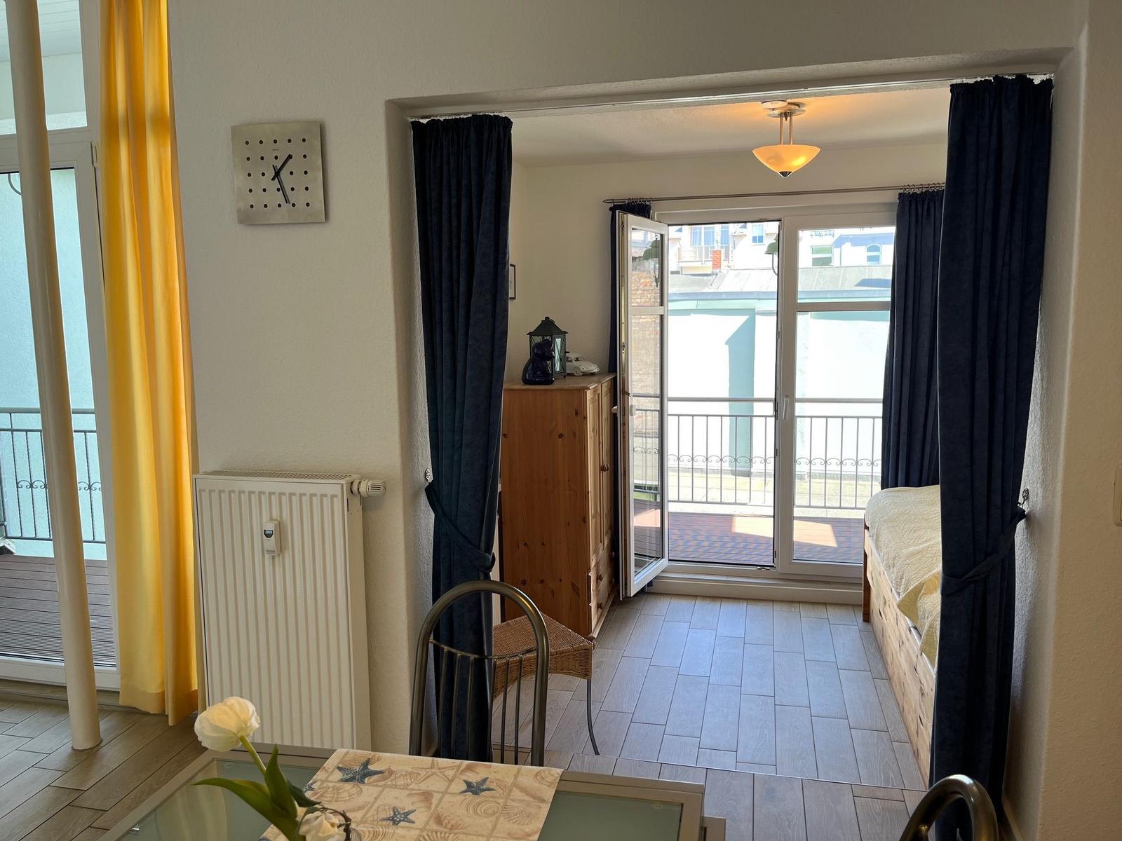 Zimmer mit Balkon, Holzmöbel und gelben Vorhängen. Blick auf Terrasse und Gebäude.