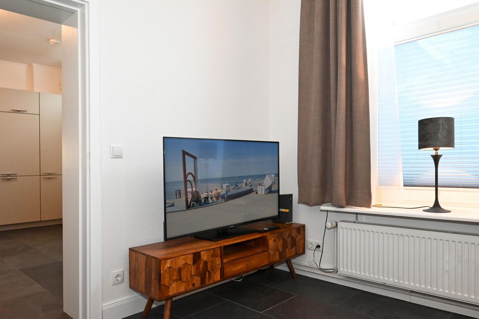 TV auf Holzboard, Fenster mit Jalousien und Vorhang, Sicht in Küche