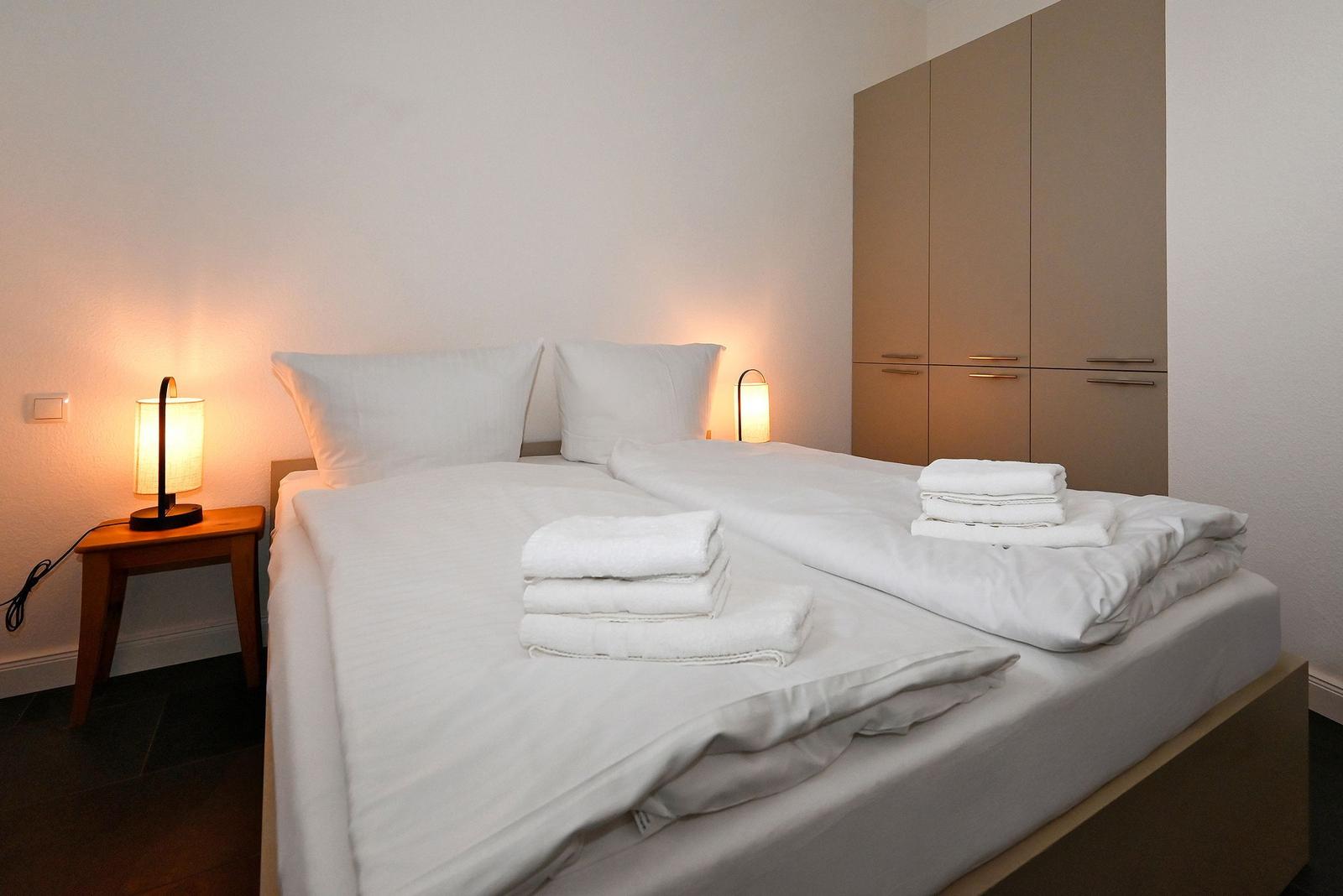 Doppelbett mit weißer Bettwäsche, zwei Nachttische mit Lampen und Kleiderschrank.