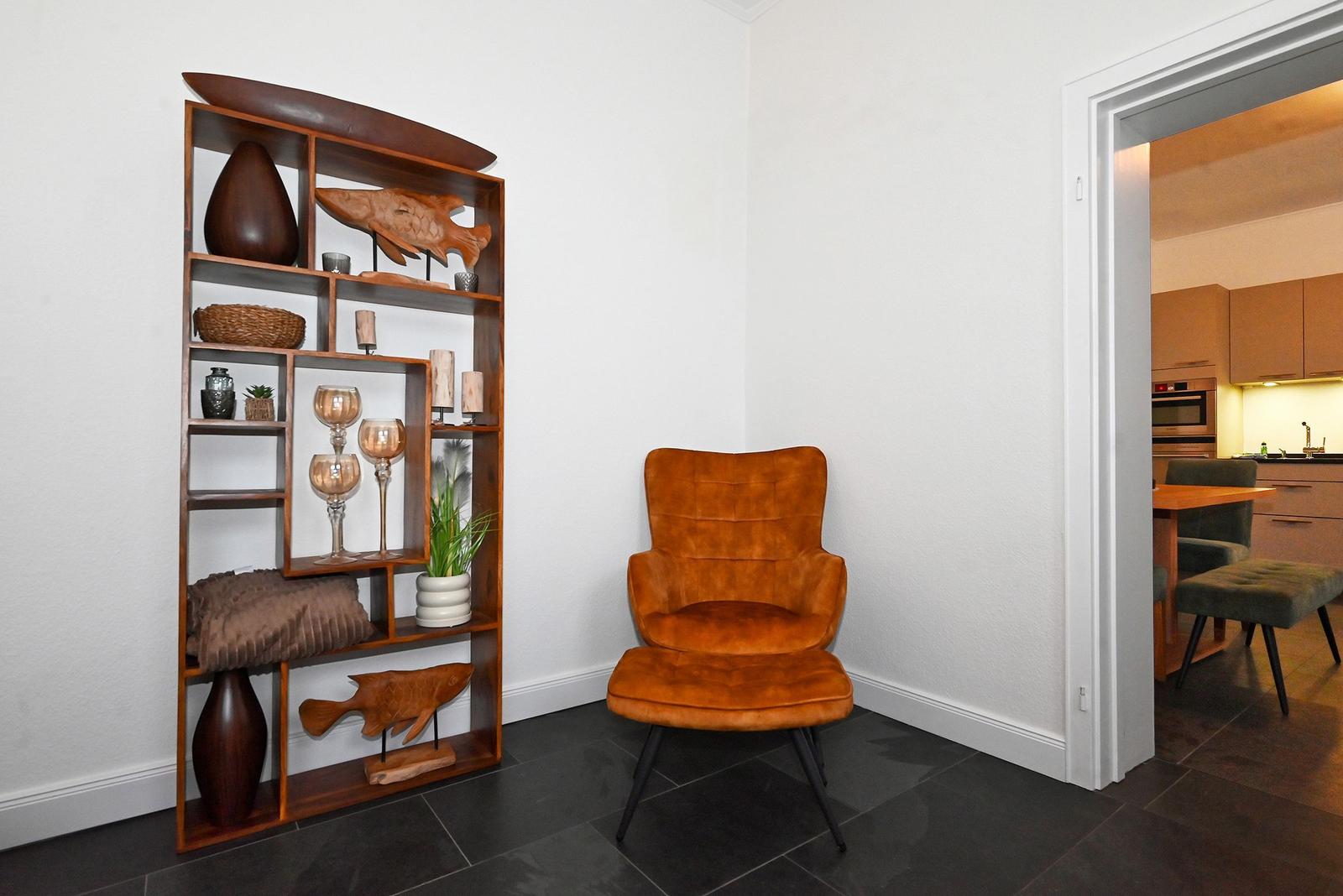 Eckzimmer mit brauner Sitzgelegenheit und Holzregal mit Dekoration. Durchgang zur Küche.