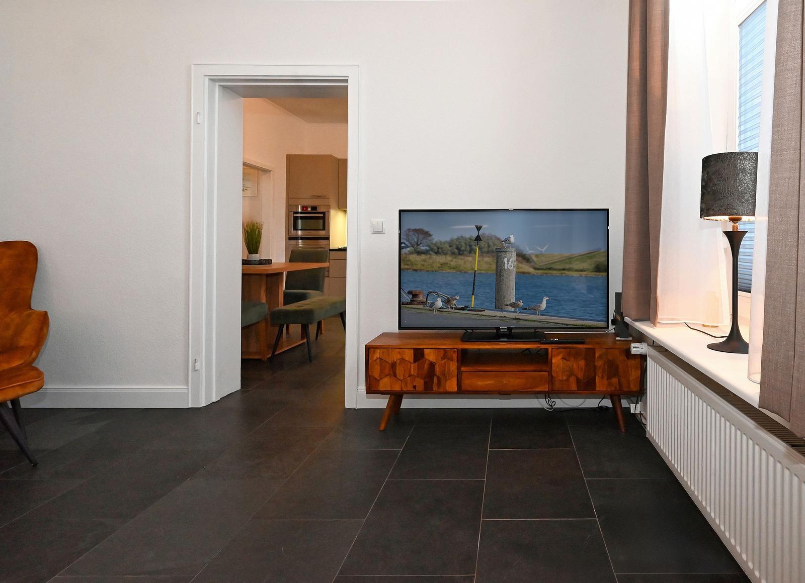 Wohnzimmer mit TV, Holzboard und Blick in die Küche.