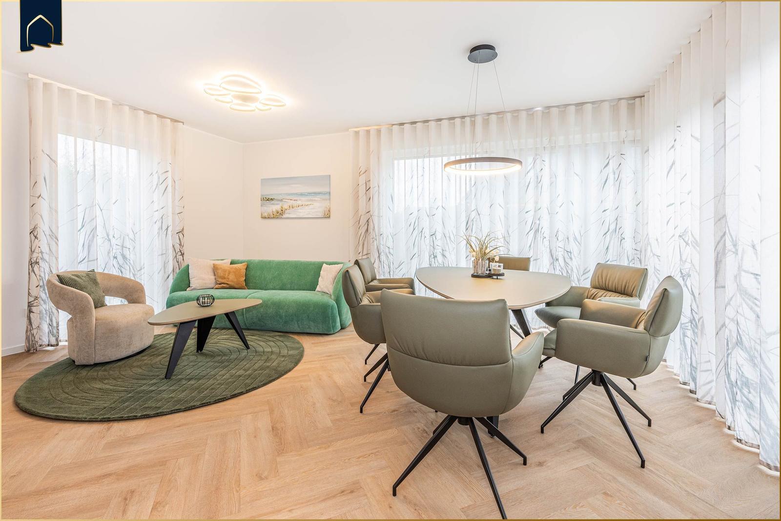 Wohn- und Essbereich mit grünem Sofa, runden Tisch und Stühlen, Holzboden und Fenstern mit Vorhängen.