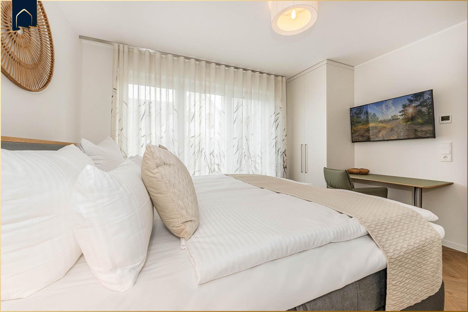 Doppelbett mit weißen Laken, Schreibtisch und TV an der Wand.