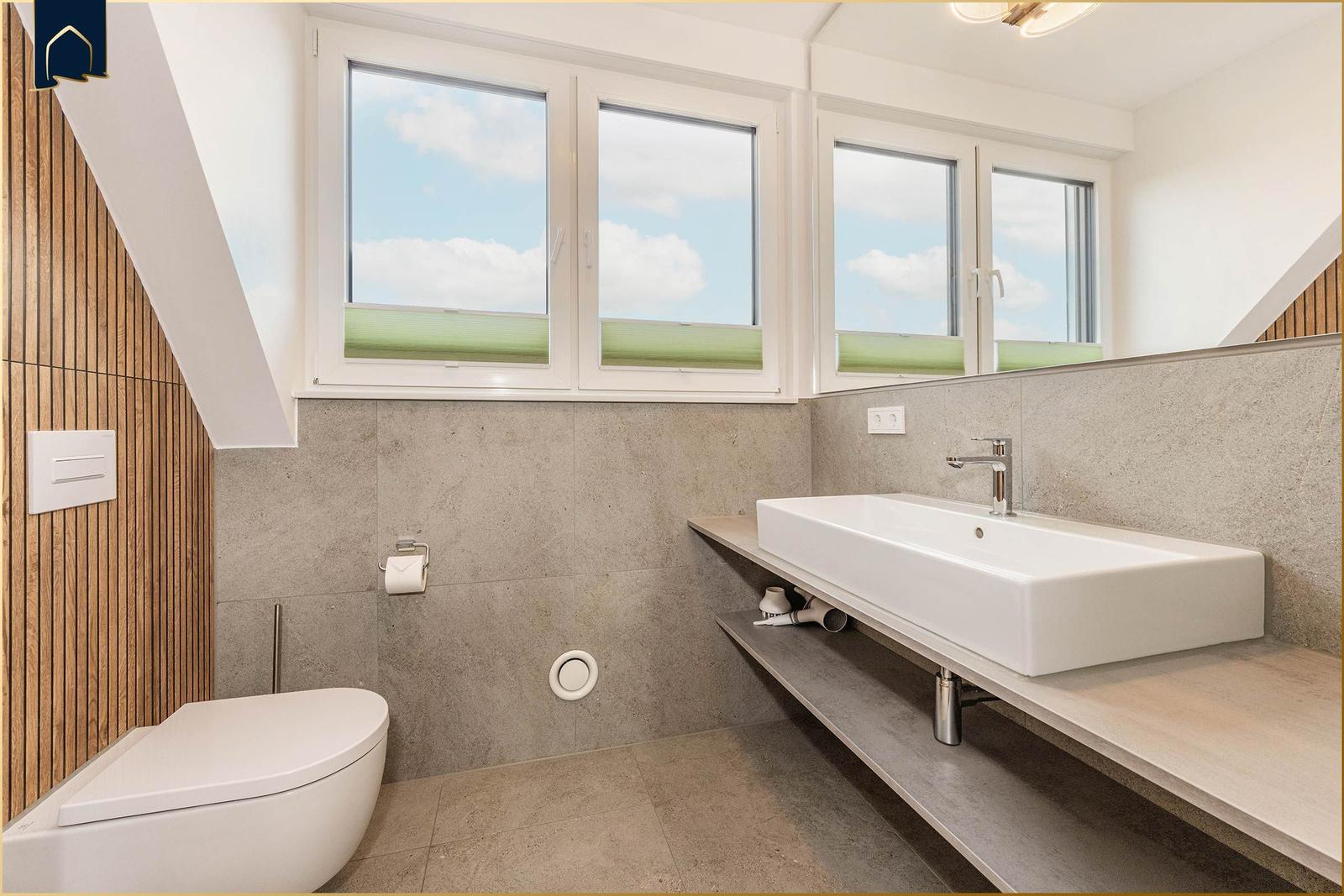 Badezimmer mit Waschtisch, Toilette und Fenstern. Holzwand und Betonfliesen.