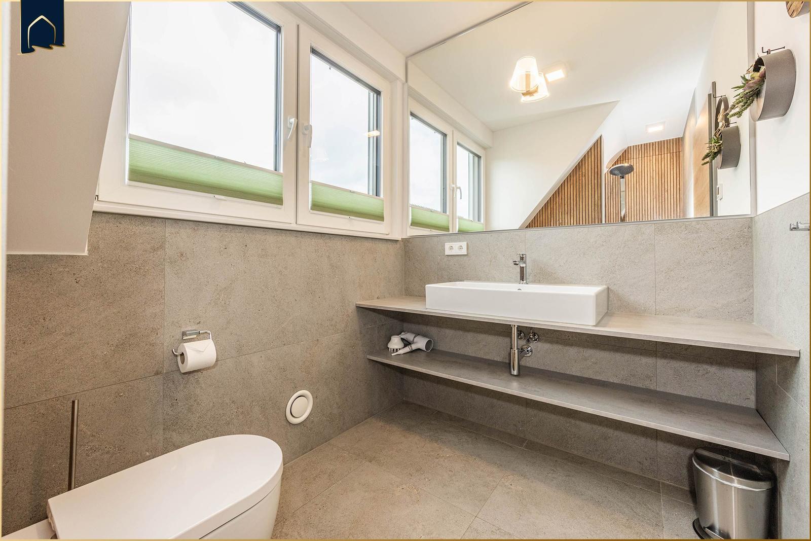 Badezimmer mit Waschtisch, Spiegel, Toilette und Fenstern.