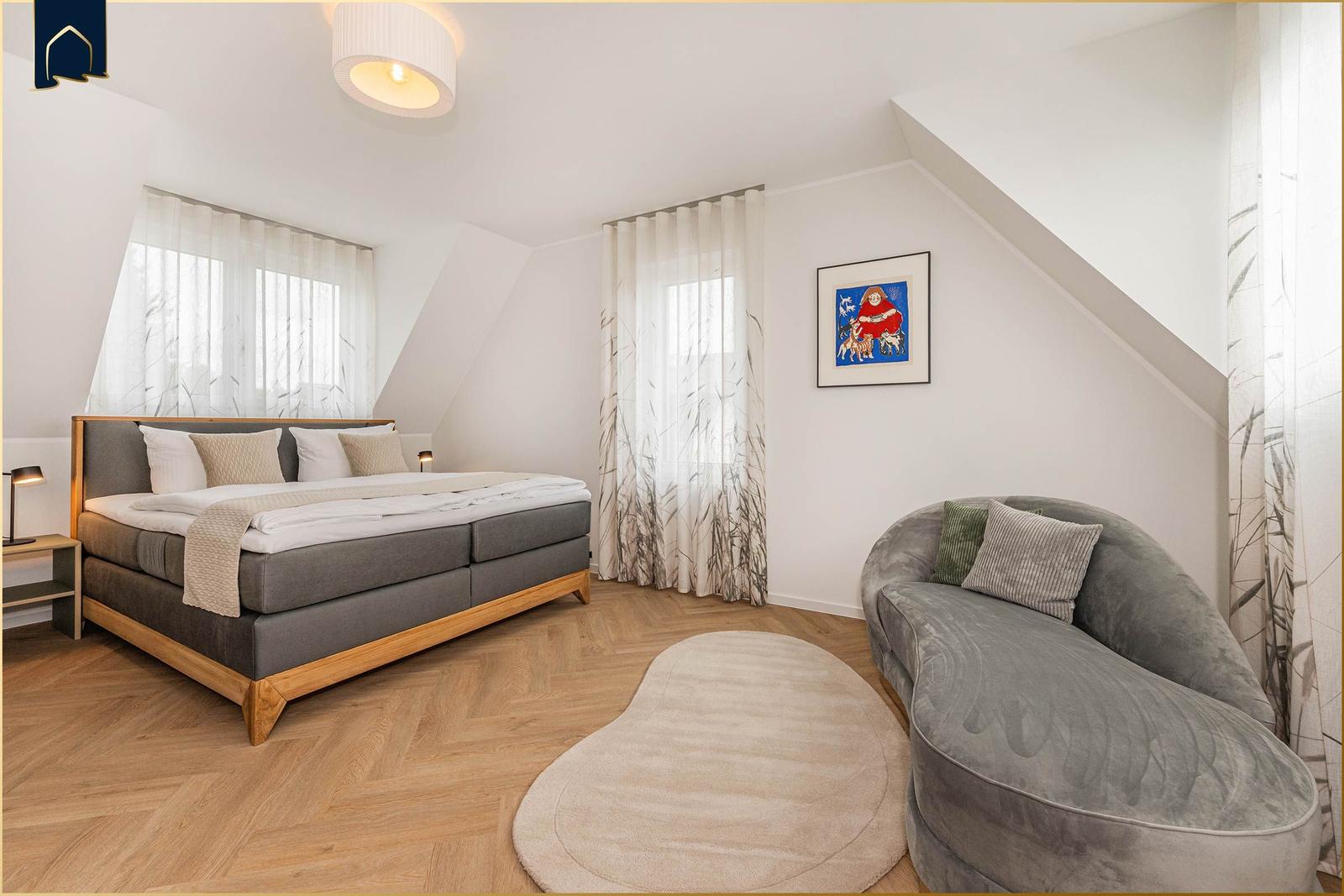 Schlafzimmer mit Doppelbett, Sessel und Holzboden. Dachschrägen und Fenster mit Vorhängen.