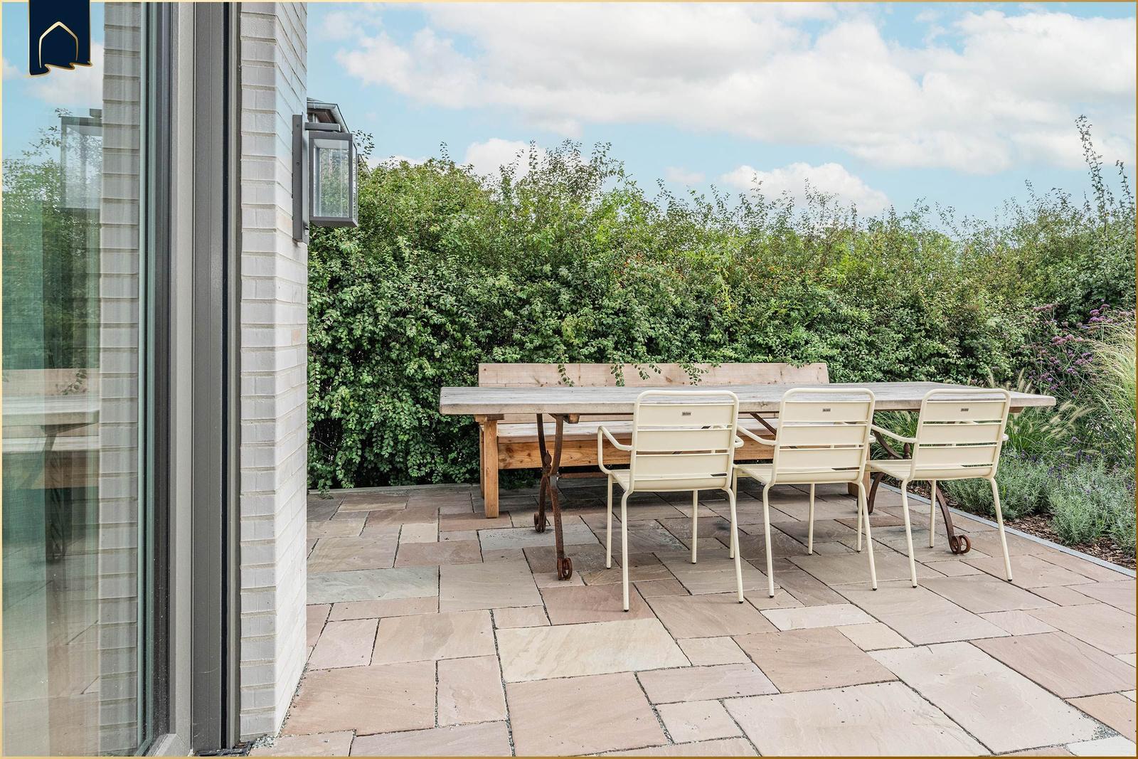 Terrasse mit Holztisch, drei Stühlen und Sitzbank. Hintergrund: grüne Hecke und blauer Himmel.