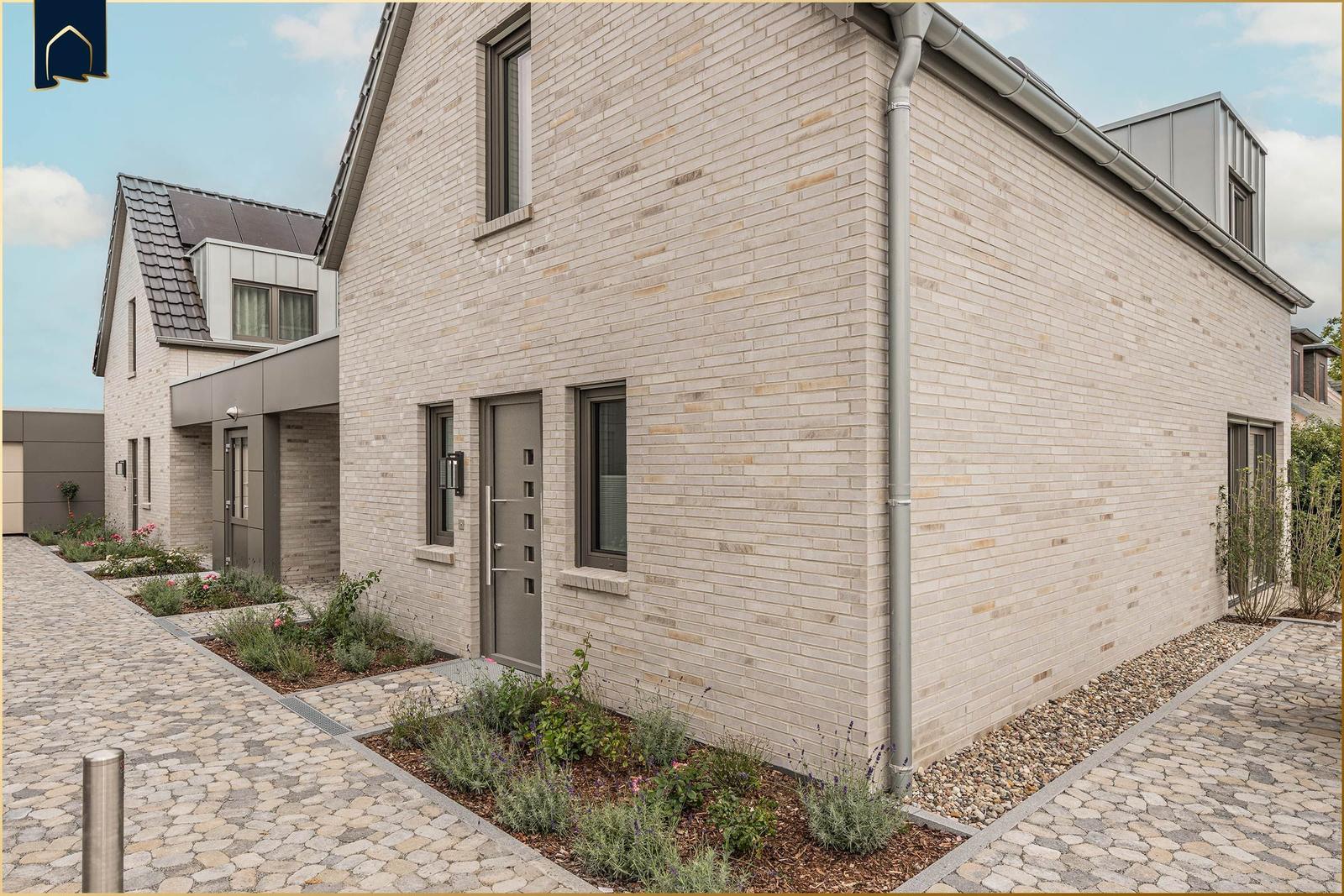 Moderne Villa mit Steinmauer, grauer Tür und gepflastertem Hof. Gartenbereich mit Pflanzen.