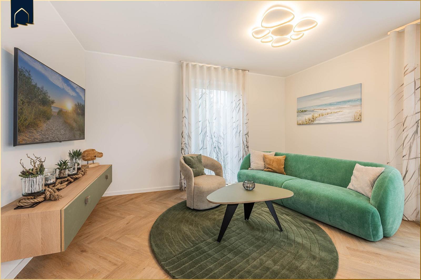 Ein Wohnzimmer mit grünem Sofa, runden Teppich und Wandbild. Fernseher und Sessel sind vorhanden.