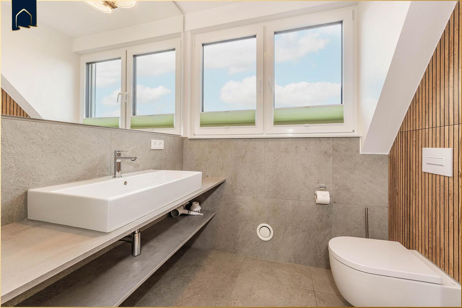 Badezimmer mit Waschbecken, Toilette und Fenstern. Wandverkleidung aus Holz und Beton.