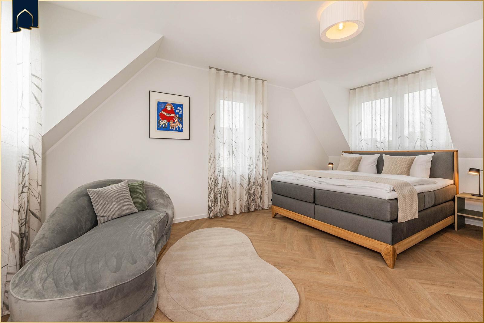 Schlafzimmer mit Doppelbett, Sofa und Holzboden. Fenster mit Vorhängen.