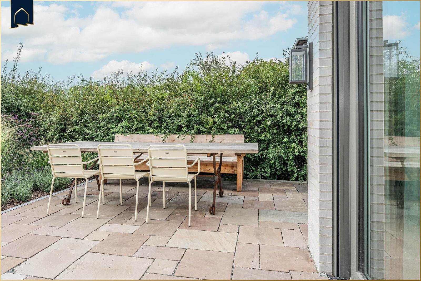 Terrasse mit Tisch, drei Stühlen und Bank. Hintergrund: grüne Hecke und Himmel.
