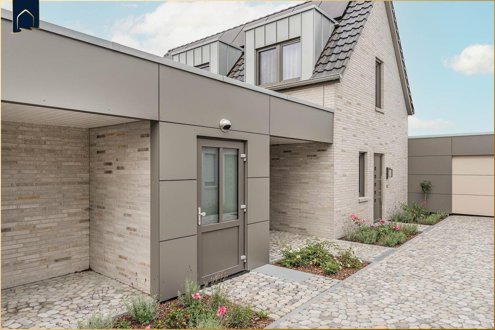 Moderne Villa mit Steinmauer, Glasfront und gepflastertem Hof. Eingangstür und Gartenbereich sichtbar.