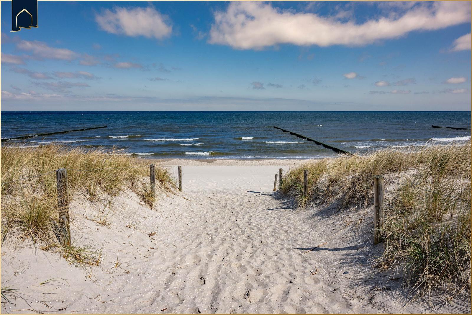 Sandweg führt von Dünen zur Ostsee mit Wellen und Holzpfosten.