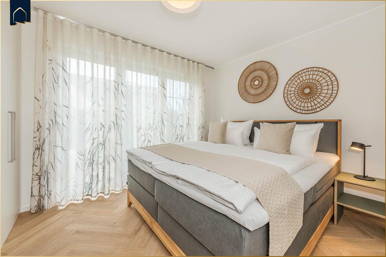 Schlafzimmer mit Doppelbett, Holzfußboden und weißen Wänden. Vorhänge mit Pflanzenmotiv und Wanddekoration.