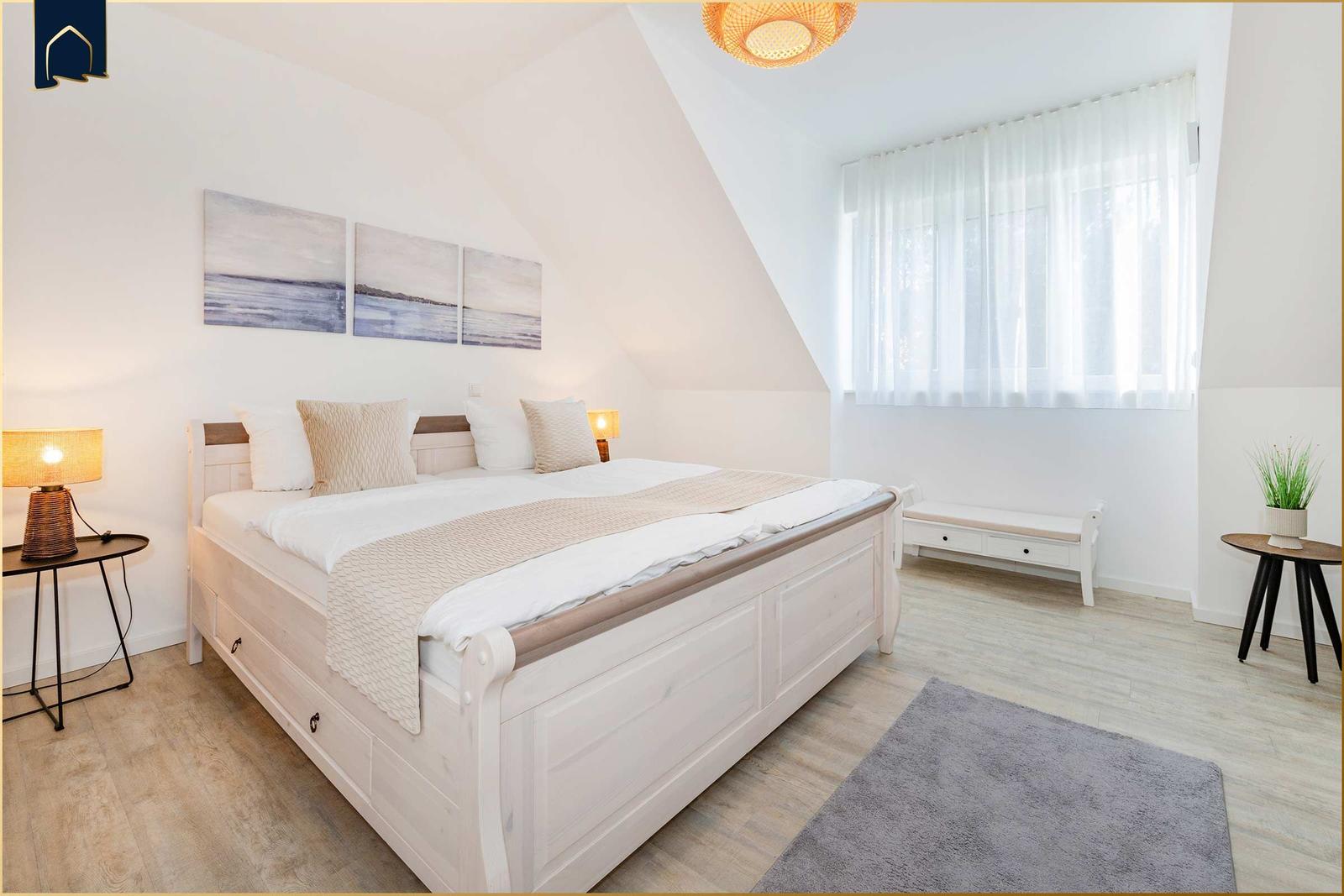 Schlafzimmer mit Doppelbett, Fenster, Nachttische und Wandbildern.