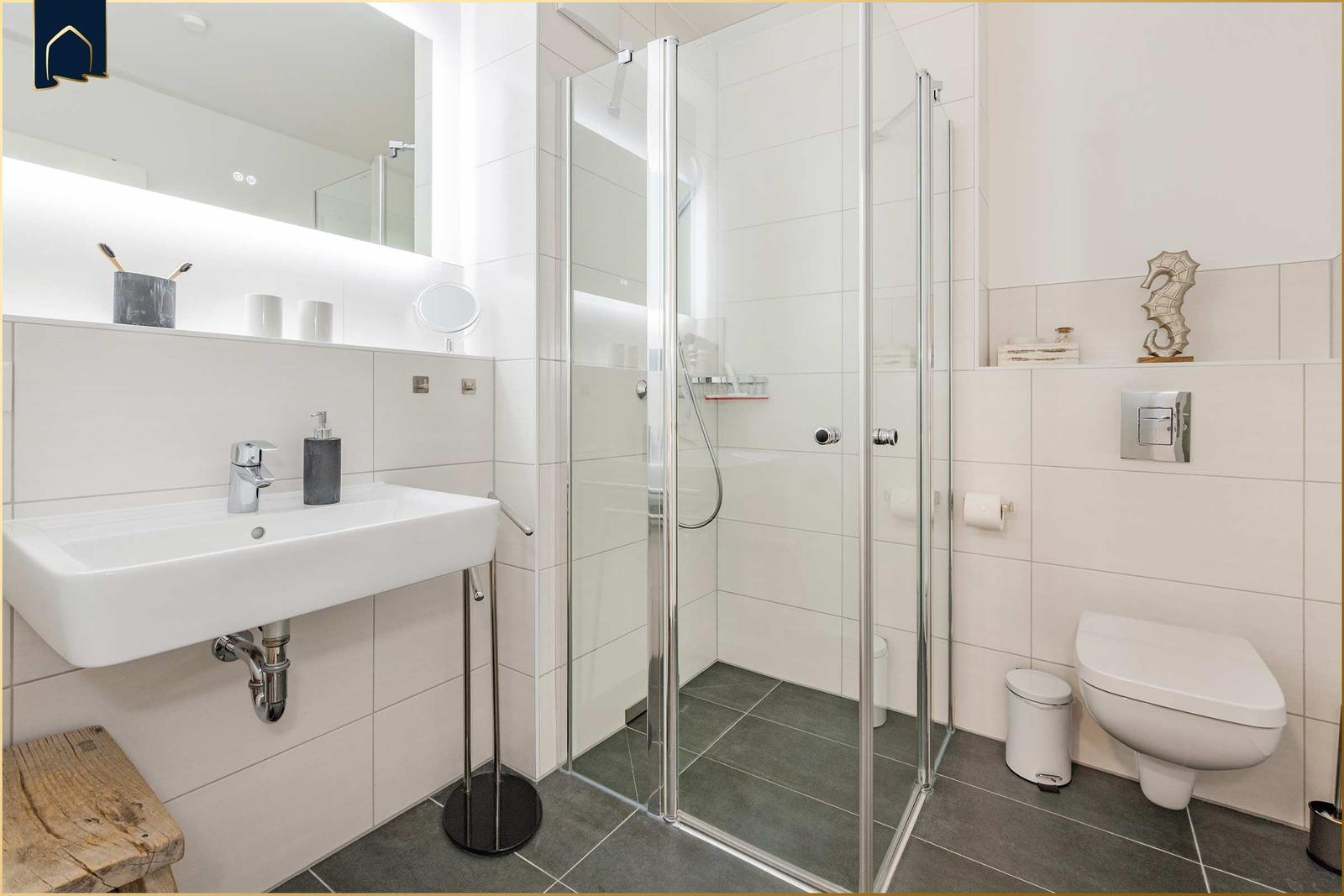 Badezimmer mit Dusche, Waschbecken und Toilette. Weiße Fliesen und graue Bodenfliesen.