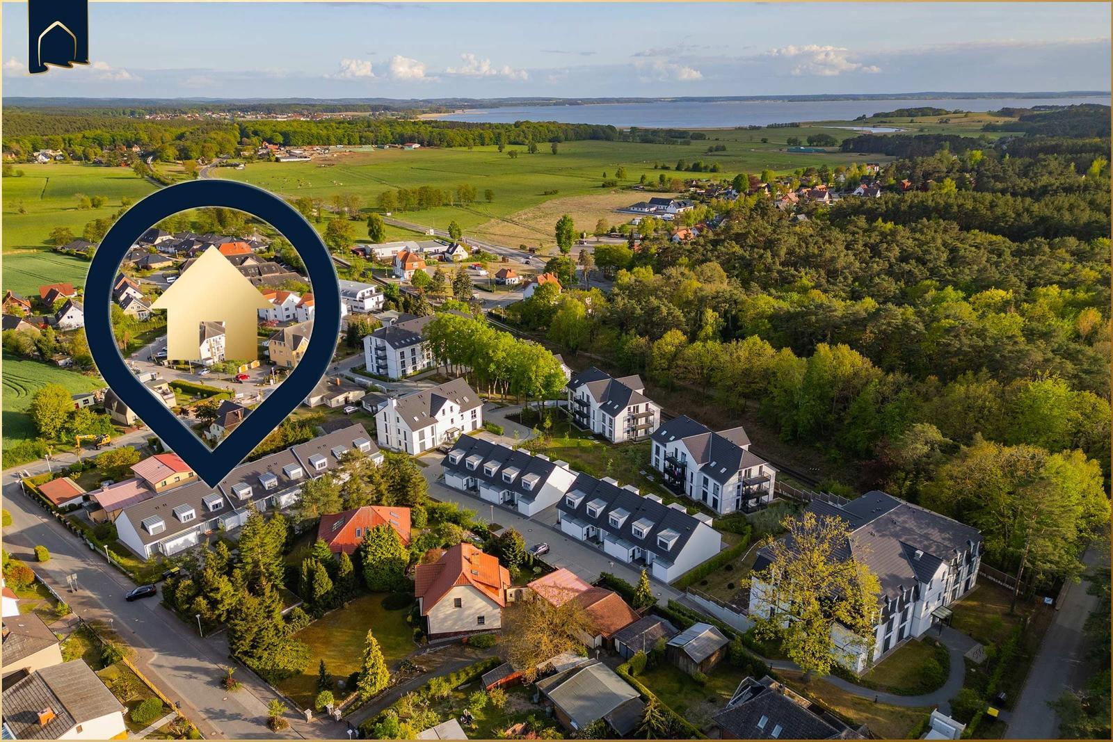Luftbild eines Dorfes mit Häusern, Wiesen und einem See im Hintergrund.