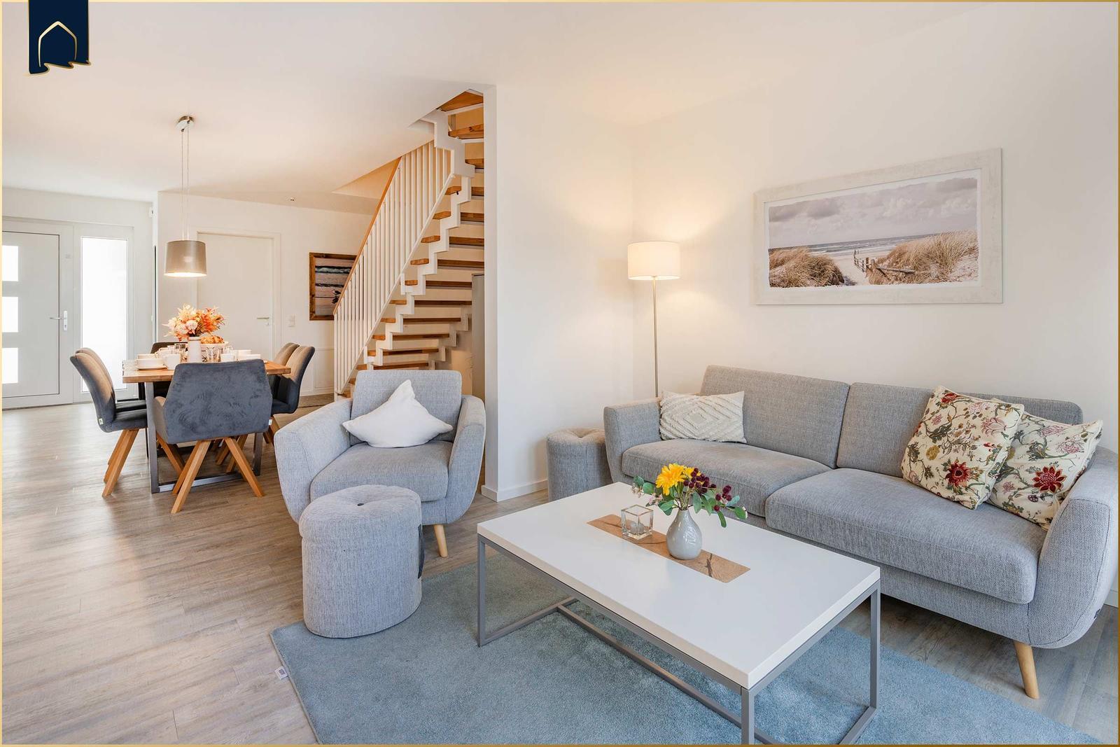 Wohn- und Essbereich mit Sofa, Sessel und Treppe. Wandbild zeigt Strand.