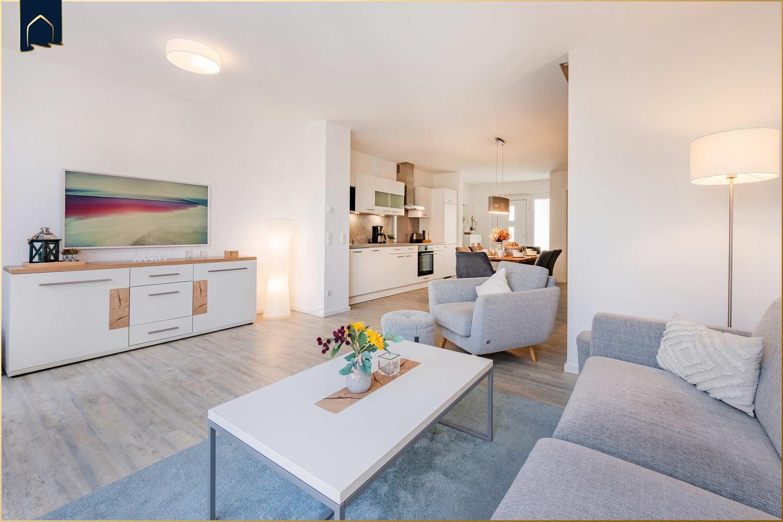 Wohnbereich mit Sofa, Sessel und Esstisch. Küche im Hintergrund. Holzboden und weiße Wände.