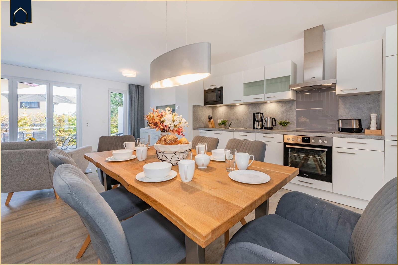 Essbereich mit Holztisch und Stühlen. Küche mit weißen Schränken und Edelstahlgeräten.