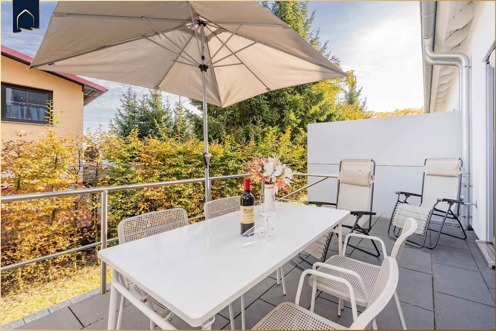 Terrasse mit Tisch, Stühlen und Sonnenschirm. Hintergrund: Bäume und ein weißer Zaun.