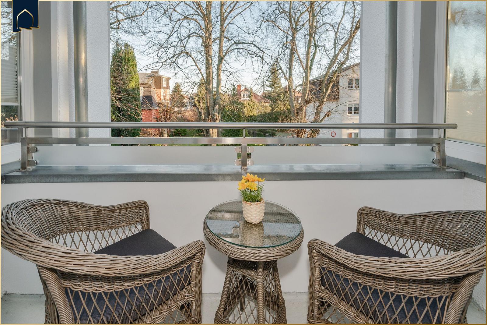 Balkon mit zwei Stühlen und Tisch. Gelbe Blumen auf Tisch. Blick auf Bäume und Gebäude.