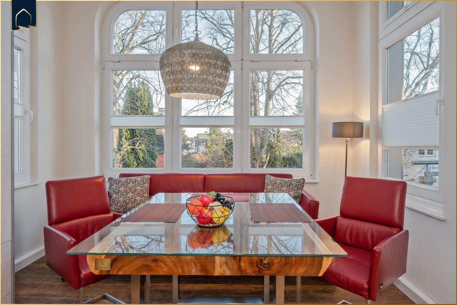 Esszimmer mit rotem Ledersofa, Glas-Tisch und großen Fenstern.