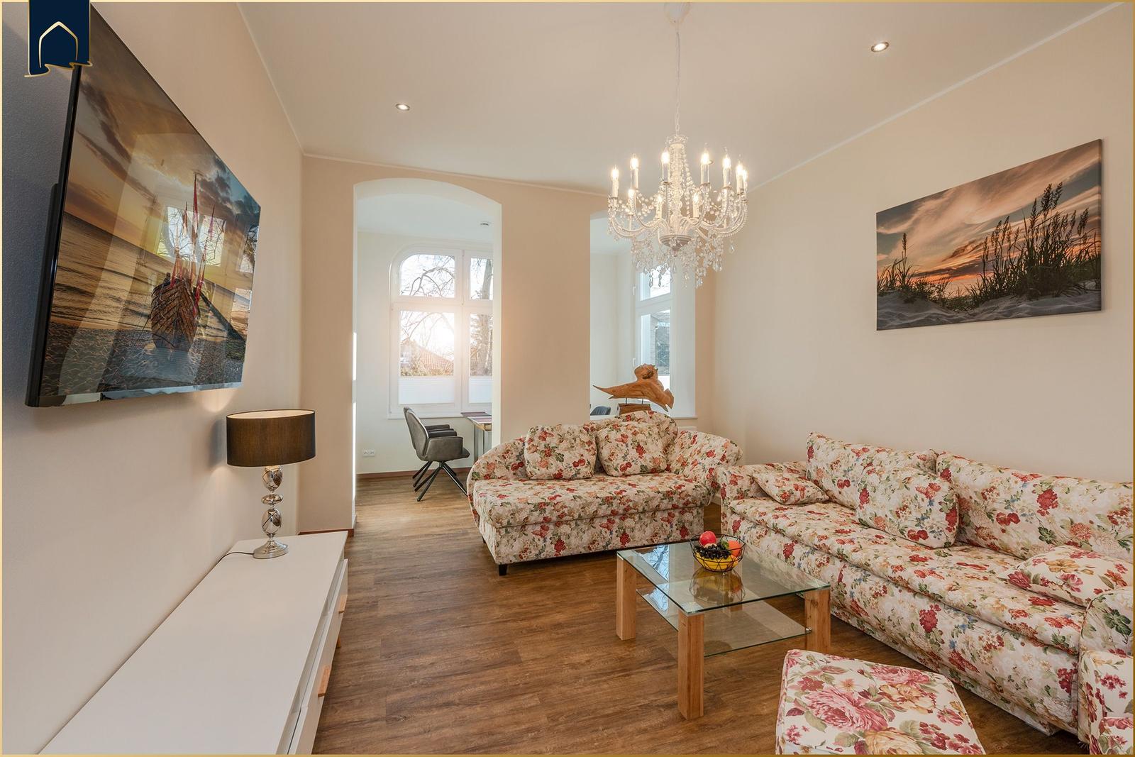 Wohnzimmer mit floralen Sofas, Glas-Tisch und Kronleuchter. Wandbild und Fernseher an der Wand.