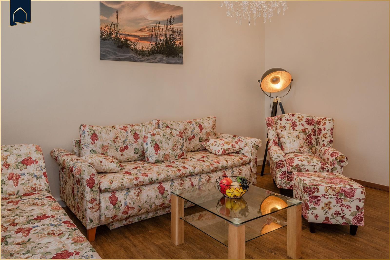 Wohnzimmer mit floralen Sitzmöbeln, Glas-Tisch und Wandbild.
