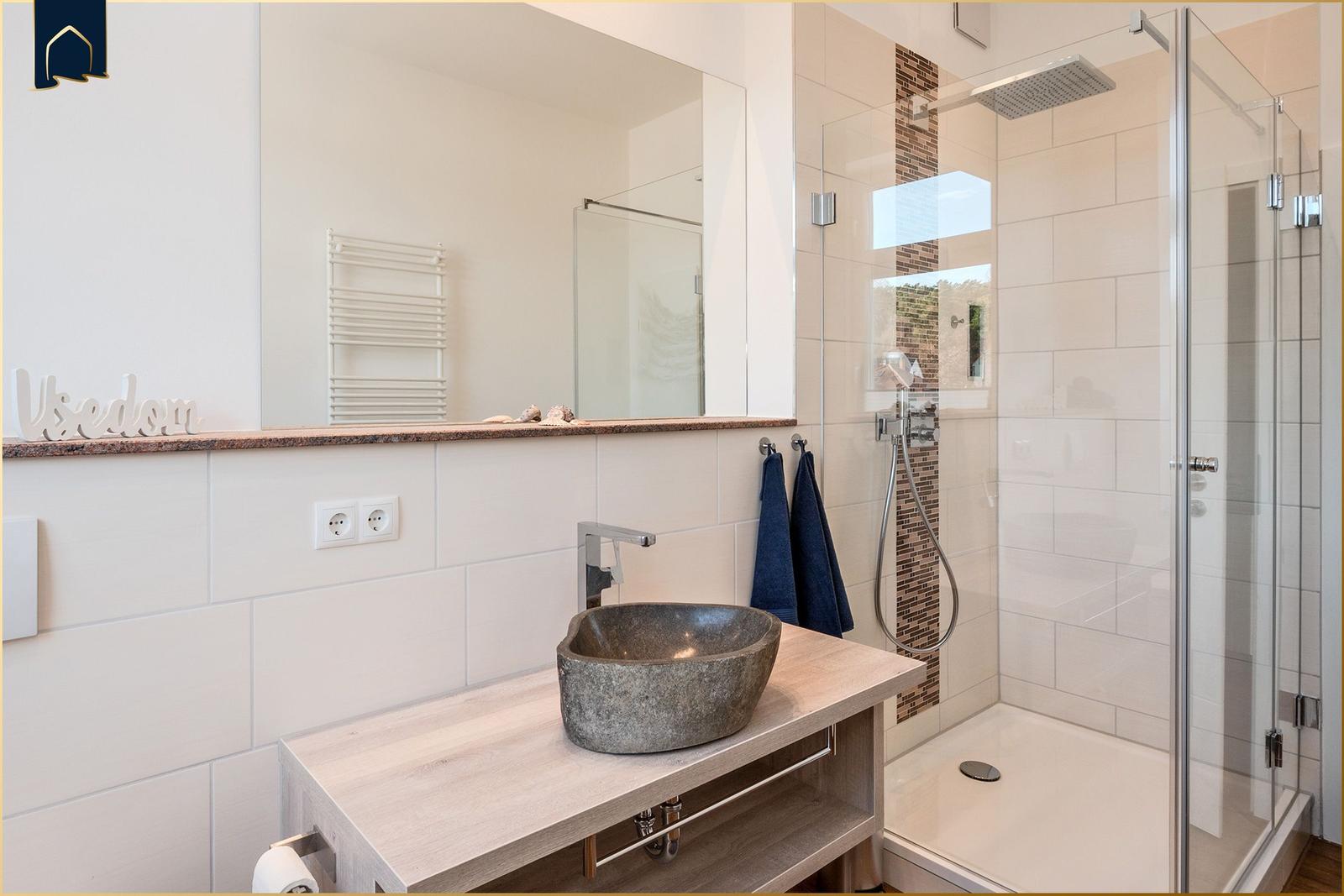 Bad mit Dusche, Steinwaschbecken und Spiegelschrank. Handtuchhalter und Steckdosen sind vorhanden.