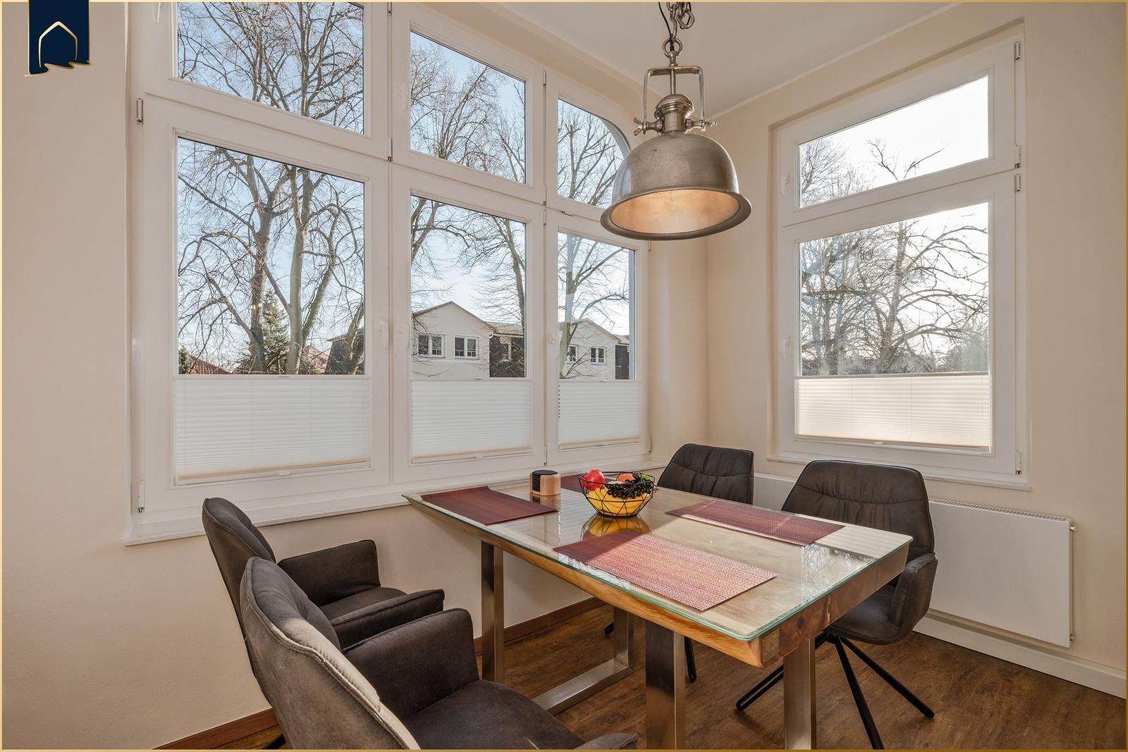 Esszimmer mit Glas-Tisch, Stühlen und großen Fenstern. Draußen sind Bäume und Häuser sichtbar.